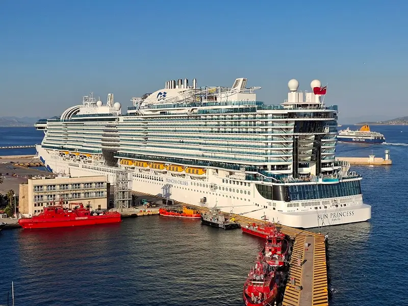 The Sun Princess cruise ship by Princess Cruises docked at Piraeus Harbour, Greece, surrounded by smaller red tugboats and port vessels with calm blue waters and coastal hills in the background.