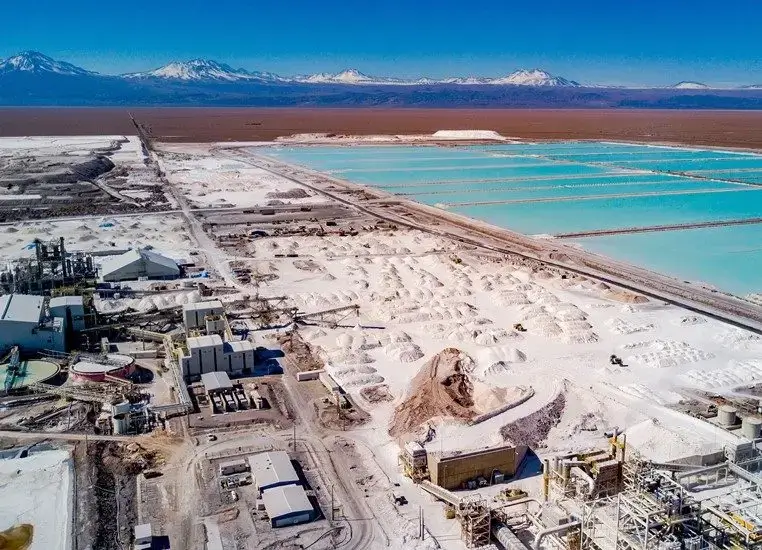 Aerial view of the SQM lithium mining operation at Salar de Atacama, Chile, showing large evaporation ponds with turquoise brine, white salt flats, industrial processing facilities, and snow-capped Andean mountains in the background.